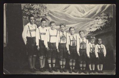Front shows 8 boys of varying ages in traditional Bavarian Lederhosen (leather shorts) with knee-high socks, white shirts, and suspenders, lined up by height against a painted mountain backdrop. They are identified as the Bohren brothers.