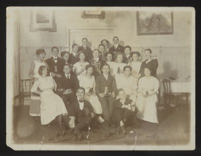 Front shows a large group of approximately 20 young people at a formal dance or social event, some holding gingerbread hearts. Taken in a room with landscape paintings on the walls. The group appears to be from the 1911-1912 winter dance season.