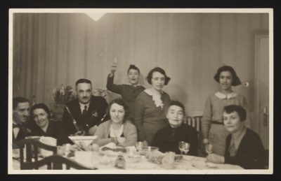 Front shows a family dinner gathering with 9 people around a table (6 adults seated, 2 young women and a boy standing behind). Glasses and dishes on the table suggest a celebration. Location: Munich, February 1932.
